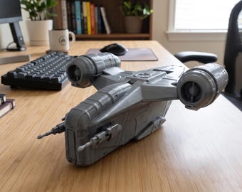 May include: A gray plastic model of a spaceship featuring two large cylindrical engines on the sides, a central cockpit section, and two forward-facing cannons. The structure displays detailed paneling and mechanical components across its surface.
