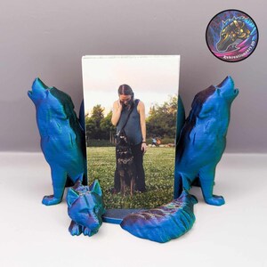 May include: A 3D printed photo frame with two blue wolves on either side and a smaller wolf in the front. The frame is holding a photo of a person with a dog.