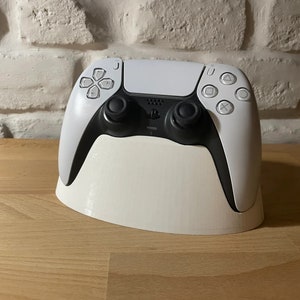 May include: A white PlayStation 5 controller sits on a white stand. The controller has black buttons and analogue sticks.