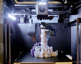 May include: A 3D printer printing a white plastic model of a dragon attacking a tower. The dragon is green and blue and has a detailed design. The tower is white and has a detailed design. The printer is a white and gray machine with a black base.