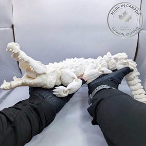 Puede incluir: Una figura de caimán articulada de color blanco roto, impresa en 3D, con un cuerpo texturizado y segmentado y la boca abierta mostrando dientes afilados. La figura es sostenida por dos manos con guantes negros, con un logo Made in Canada visible.