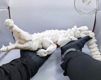 May include: An off-white, articulated 3D-printed alligator figurine with a textured, segmented body and an open mouth showing sharp teeth. The figure is held by two hands wearing black textured gloves, with a circular Made in Canada logo visible.