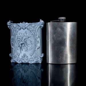 May include: A silver flask with a brushed finish and a silver cigarette case with an ornate design featuring a woman in a flowing dress.