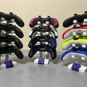 Peut inclure: Un support blanc à quatre niveaux contient neuf manettes de jeu vidéo. Les manettes sont de différentes couleurs, notamment noir, blanc, rouge, bleu, vert et jaune. Le support a une base violette.