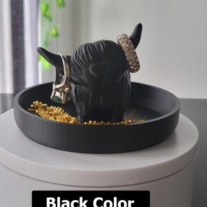 以下が含まれることがあります： 丸いベースの黒いハイランド牛型のジュエリーホルダー。ホルダーには小物のためのトレイとリングを保持するための角があります。「Black Color」というテキストが画像の下部に表示されています。