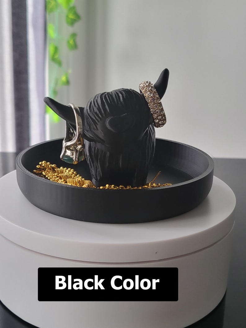 以下が含まれることがあります： 丸いベースの黒いハイランド牛型のジュエリーホルダー。ホルダーには小物のためのトレイとリングを保持するための角があります。「Black Color」というテキストが画像の下部に表示されています。