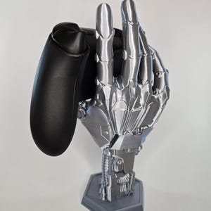 Könnte beinhalten: Eine silberne Roboterhand hält einen schwarzen Videospielcontroller. Die Hand befindet sich auf einem sechseckigen Sockel.
