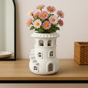 May include: A white ceramic lighthouse-shaped vase filled with pink and yellow daisies. The vase has arched windows and a spiral staircase design. The flowers are in full bloom. The vase is on a wooden surface.