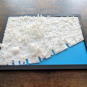 以下が含まれることがあります： 黒い枠で囲まれた、青い背景の白い3Dプリントされた都市の街並みモデル。モデルは詳細で、建物、通り、川が含まれています。