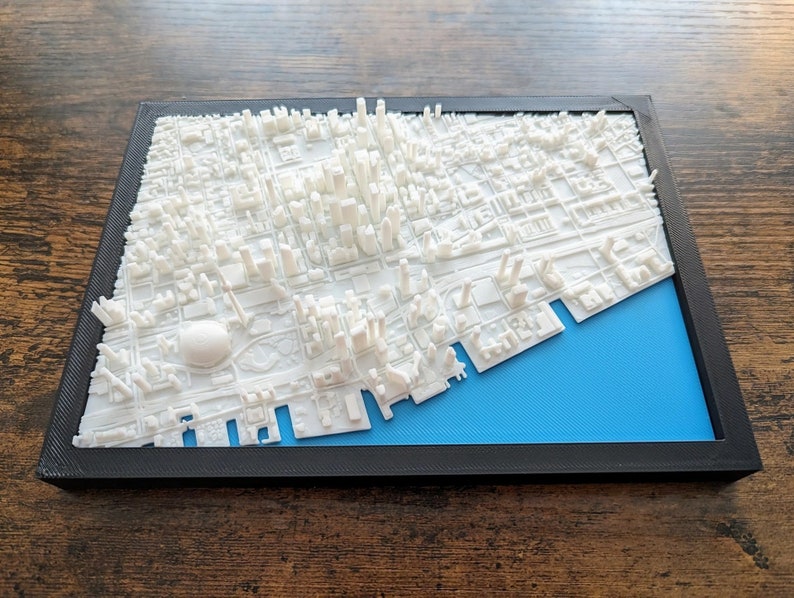 以下が含まれることがあります： 黒い枠で囲まれた、青い背景の白い3Dプリントされた都市の街並みモデル。モデルは詳細で、建物、通り、川が含まれています。