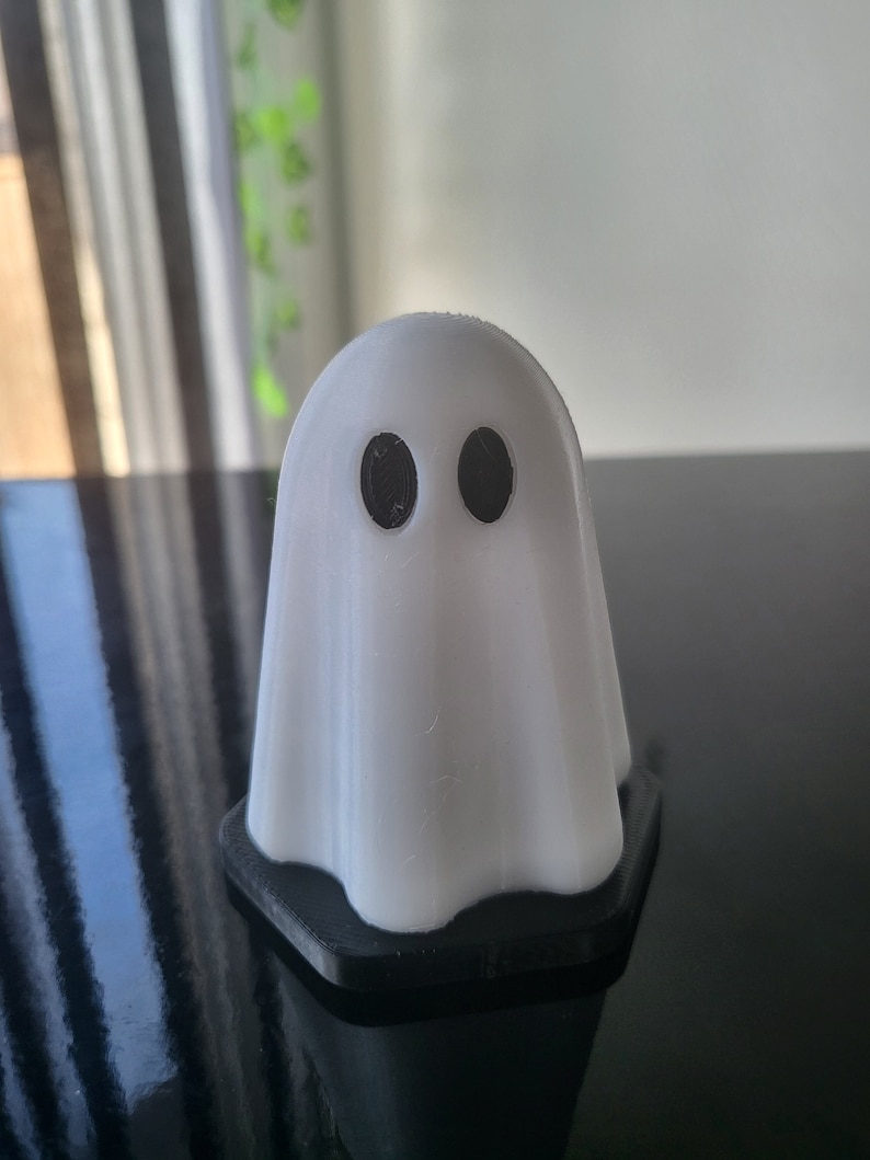 Puede incluir: Una figurita de fantasma blanca con ojos negros, de pie sobre una base negra. El fantasma tiene la forma de un fantasma tradicional con una cabeza redonda y una s&aacute;bana que fluye.