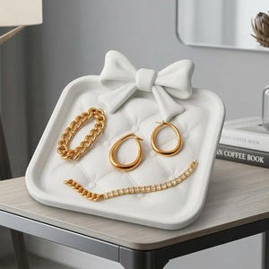 Op de afbeelding: Een witte sieraden tray in de vorm van een strik, met een gewatteerd oppervlak, toont gouden sieraden. De sieraden omvatten een kettingarmband, een tennisarmband en twee paar oorbellen. De tray staat op een lichtgekleurde tafel.