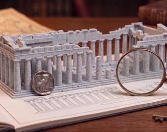 May include: A light blue, speckled 3D-printed model of the Parthenon temple featuring a colonnade, positioned on an open book with architectural drawings. A silver owl coin, a brass magnifying glass, a metal thimble, and a small key are nearby.