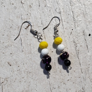 May include: A pair of dangle earrings with silver-colored hooks. Each earring features a series of beads in yellow, white, black, and purple. The beads are various shapes and sizes, creating a unique and colorful accessory.