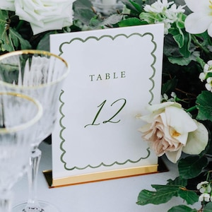 May include: A white table number card with green text that reads "TABLE 12" sits on a white table with a centerpiece of white flowers and greenery. Two clear glass wine glasses with gold rims are on the table.