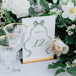 Könnte beinhalten: Ein weißer Tisch mit einem grünen und weißen Blumenschmuck. Eine weiße Karte mit grüner Schrift zeigt "Tisch 12".