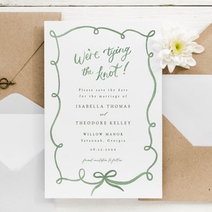 May include: Save the date card with a green calligraphy border and a bow at the bottom. The text reads "We're tying the knot!" followed by "Please save the date for the marriage of Isabella Thomas and Theodore Kelley, Willow Manor, Savannah, Georgia, 06.11.202X, formal invitation to follow."