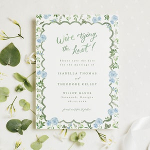 May include: A wedding invitation with a floral design. The invitation features the text "We're tying the knot!" and includes details for Isabella Thomas and Theodore Kelley. The invitation is surrounded by blue flowers and green foliage.