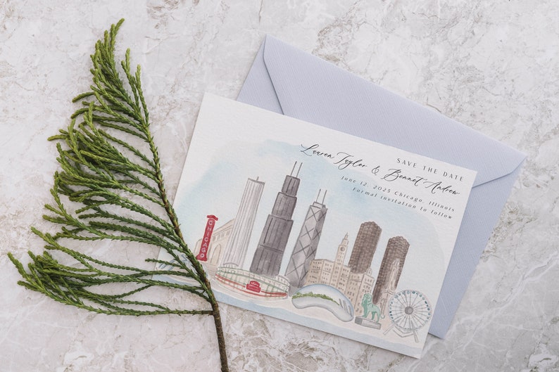 May include: A watercolor-style save-the-date card featuring a Chicago skyline illustration, with the text "Lauren Tyler & Bennet Arber" and "June 12, 2025 Chicago, Illinois." A light blue envelope is behind the card. A sprig of green foliage is in the foreground.