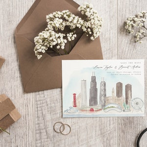 May include: A wedding invitation with a Chicago skyline illustration, accompanied by a brown envelope, gift boxes, and sprigs of white flowers. The invitation reads "Save the Date" with names and a date. Two gold rings and scissors are also present.