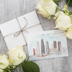 May include: A white envelope tied with twine next to a watercolor save-the-date card featuring a Chicago skyline illustration. The card includes the text "Save the Date" and names. White roses are arranged around the card.