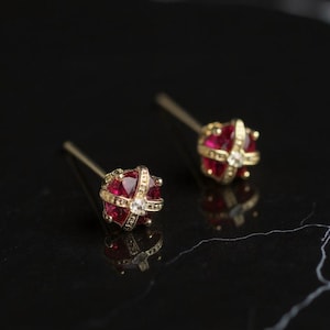 May include: A pair of gold-colored stud earrings. Each earring features a cluster of red gemstones and a central clear gemstone, set within a decorative gold frame. The earrings are displayed on a black surface.