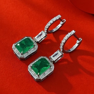 May include: A pair of silver drop earrings with emerald-cut green gemstones. The earrings have a rectangular emerald-cut green center stone, surrounded by smaller clear stones. The hoops are also encrusted with clear stones, set against a red background.