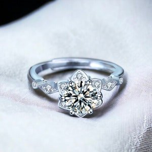 Könnte beinhalten: Ein silberner Ring mit einem großen, runden, klaren Edelstein, der von kleineren, klaren Edelsteinen in Blütenform umgeben ist.