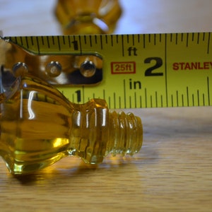May include: A close-up of a clear amber glass knob with a threaded stem. The knob is being measured with a yellow tape measure that reads "25ft" and "STANLEY".