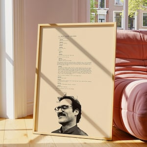 May include: A framed poster featuring a black and white portrait of a man with glasses and a mustache, with script text above. The poster is in a light wooden frame and leans against a wall. The background includes a window and a pink sofa.