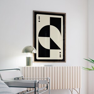 May include: A black and white geometric print of the Queen of Spades playing card in a wooden frame. The card is hanging on a white wall above a light brown cabinet with a gold lamp on top.