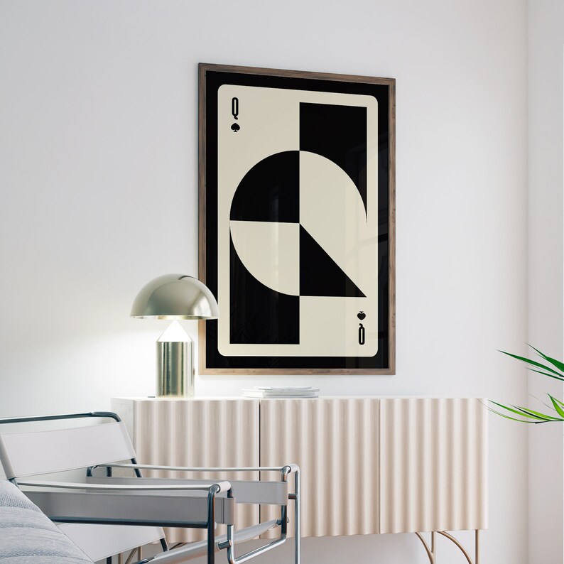 May include: A black and white geometric print of the Queen of Spades playing card in a wooden frame. The card is hanging on a white wall above a light brown cabinet with a gold lamp on top.