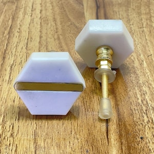 May include: Two white hexagonal cabinet knobs with a gold accent stripe. One knob has a gold screw and a clear plastic base.
