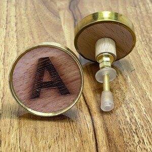 Puede incluir: Dos pomos de madera con ribete de metal dorado. Un pomo presenta la letra "A" en letras negras.