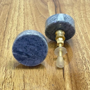 Könnte beinhalten: Zwei runde graue Marmorknopfknöpfe mit goldfarbener Metallbeschläge. Die Knöpfe befinden sich auf einer Holzoberfläche.