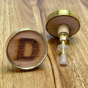 Könnte beinhalten: Zwei Holzknöpfe mit Messingrand. Ein Knopf zeigt den Buchstaben "D", der in das Holz gebrannt ist. Der andere Knopf hat eine Schraube aus Messing und eine transparente Kunststoffscheibe.