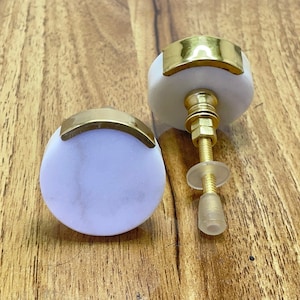 May include: Two white marble cabinet knobs with a gold metal trim. The knobs are round and have a slightly raised edge. The gold trim is a thin band that goes around the edge of the knob.