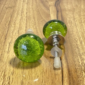Op de afbeelding: Twee groene glazen kastknoppen met een zilveren metalen basis en schroef. Het glas is getextureerd met kleine bubbels.