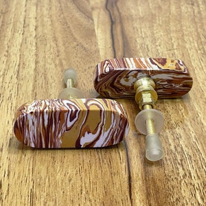 Op de afbeelding: Twee ovale kastknoppen met een bruin, wit en rood marmerpatroon. De knoppen hebben een gouden metalen schroef en een doorzichtige plastic ring.