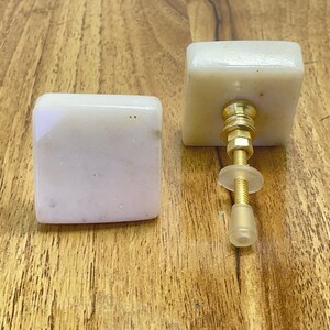 Op de afbeelding: Twee witte vierkante kastknoppen met een gouden metalen schroef en een doorzichtige kunststof basis. De knoppen hebben een gespikkelde textuur.