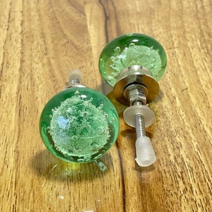 May include: Two green glass cabinet knobs with silver metal bases and screws. The knobs have a bubbly texture.