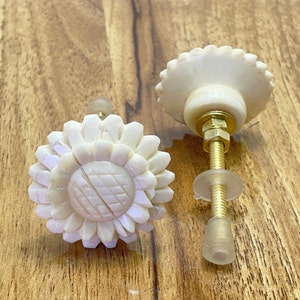May include: Two white ceramic cabinet knobs with a sunflower design. The knobs have a gold metal screw and a clear plastic washer.