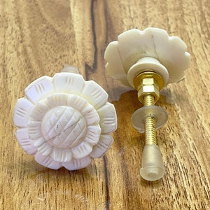 Op de afbeelding: Twee witte, gegraveerde, bloemvormige kastknoppen met gouden metalen hardware. De knoppen hebben een gedetailleerd ontwerp en zijn perfect om elke kamer een vleugje elegantie te geven.