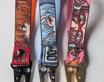 Könnte beinhalten: Drei Schlüsselanhänger mit Charakterdesigns im Anime-Stil. Die Schlüsselanhänger haben rote, blaue und rosa Bänder mit Metallverschlüssen. Jeder Riemen zeigt einen anderen Charakter mit leuchtenden Farben und kräftigen Grafiken. Die Verschlüsse sind schwarz, silber und gold.