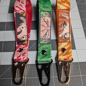 Puede incluir: Tres llaveros de tela tipo pulsera con ilustraciones de ojos de personajes de anime. El izquierdo es rojo con un remache de corazón, el central es verde con un remache de corazón negro y el derecho es naranja con un remache de corazón.