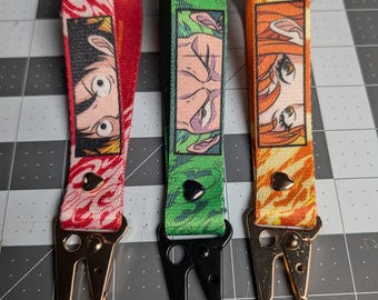 May include: Three fabric wristlet keychains featuring anime character eye illustrations. The left is red with a heart rivet, the center is green with a black heart rivet, and the right is orange with a heart rivet. Each has a metal lobster clasp.