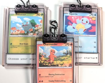 May include: Three clear acrylic card holders containing Bulbasaur, Squirtle, and Charmander trading cards. Each holder features a black pixel-art style clip with a ball chain and a white base plate printed with the text Choose your starter.