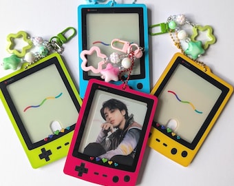 Könnte beinhalten: Eine Sammlung farbenfroher Schlüsselanhänger für Handheld-Spielkonsolen. Die Schlüsselanhänger sind in leuchtenden Farben: Grün, Blau, Gelb und Pink. Jeder Schlüsselanhänger hat einen transparenten Bildschirm und dekorative Charms.