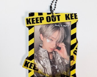 May include: A rectangular photo holder featuring a yellow and black hazard stripe border with the text KEEP OUT and a black ball chain. Inside is a portrait of a person with silver hair wearing a metallic silver jacket and the text this is mine.