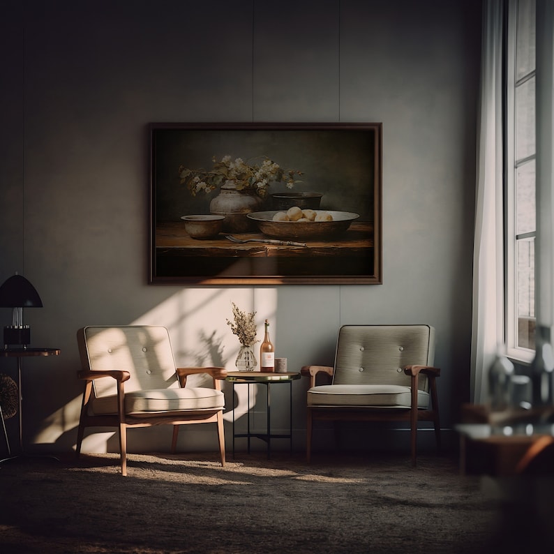 May include: A still life painting depicting a bowl of fruit, a vase of flowers, and other objects on a wooden table. The painting is framed in a dark wood frame and hangs on a grey wall. Two chairs are in the foreground, one on each side of the painting.
